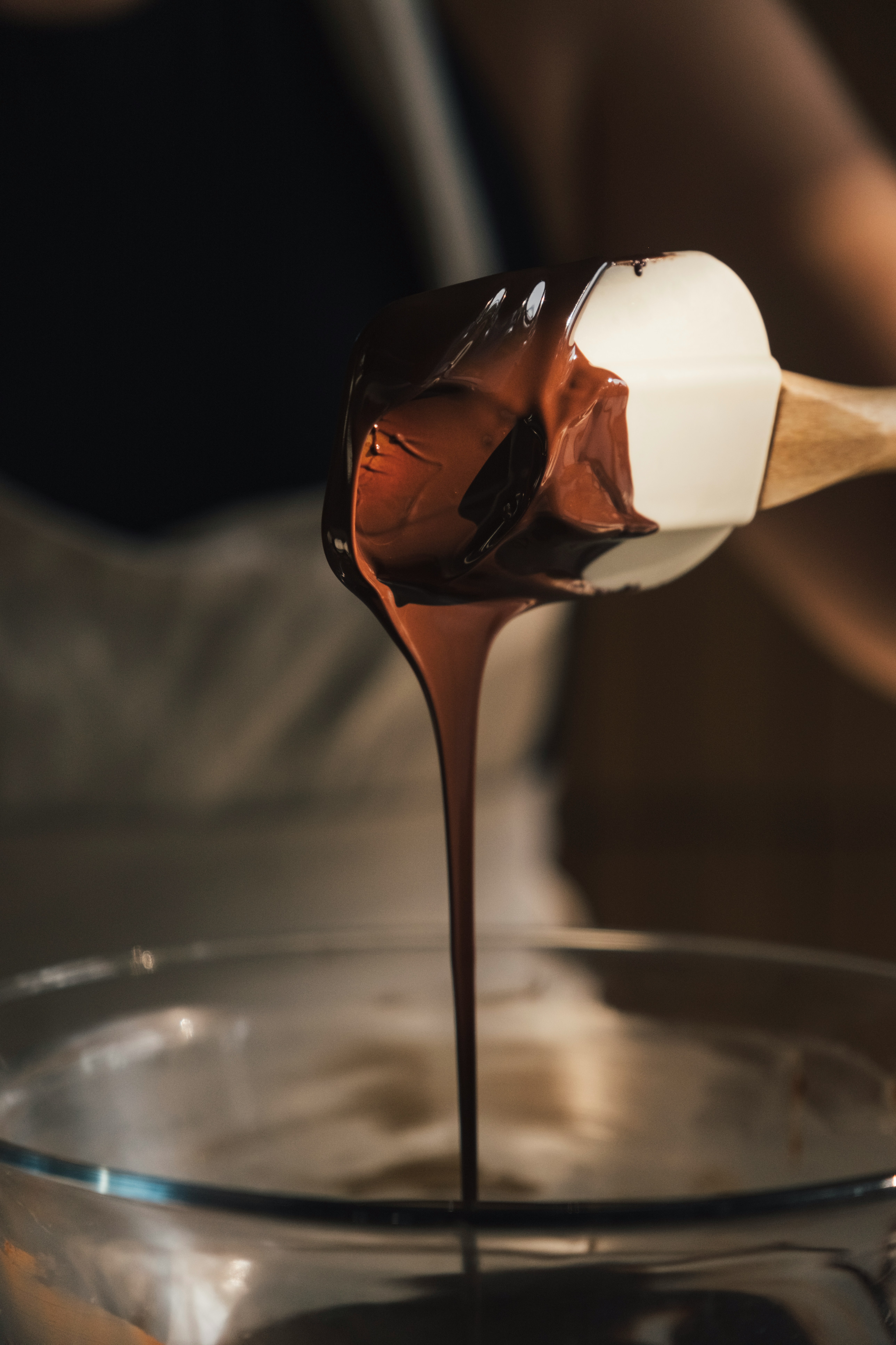 Smooth melted chocolate streaming from a spatula into a glass mixing bowl during recipe preparation.